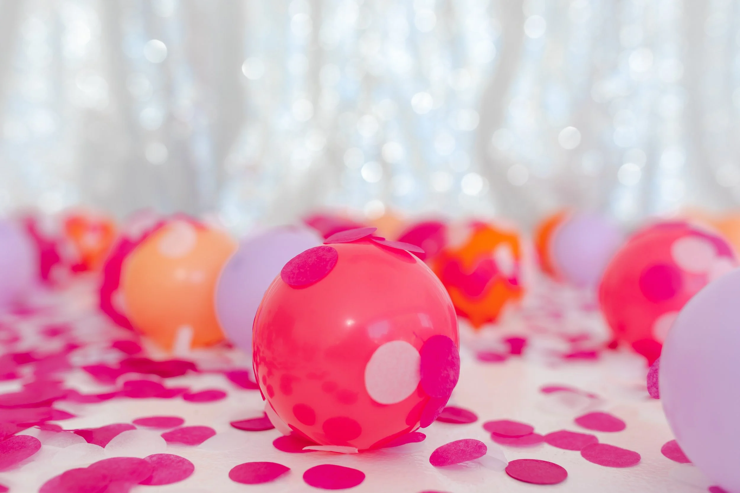 Colorful polka dot balloons and scattered confetti on a table with a blurred silver background.
