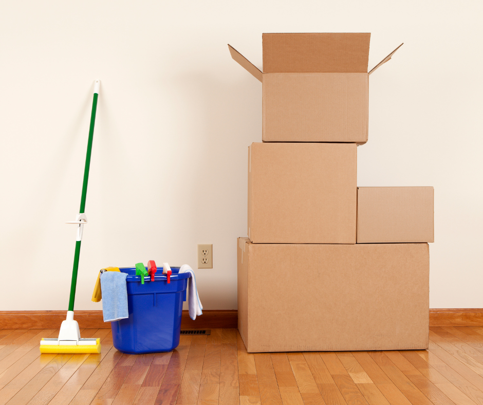 Stack of four cardboard moving boxes next to cleaning supplies, including a mop, bucket, and cleaning cloths, against a plain wall.