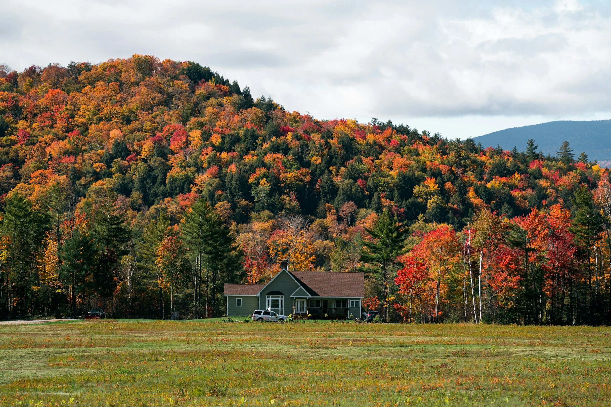 A house is situated in front of a hillside of Vermont covered with colorful autumn trees, under a cloudy sky.