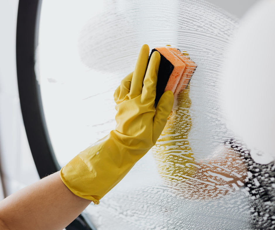 Person wearing yellow rubber gloves cleaning a glass window with a sponge, removing soap or cleaning solution.