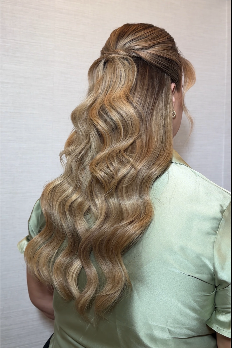 Side view of a woman with long, wavy, blonde hair styled with volume and soft curls, wearing a light green satin top.