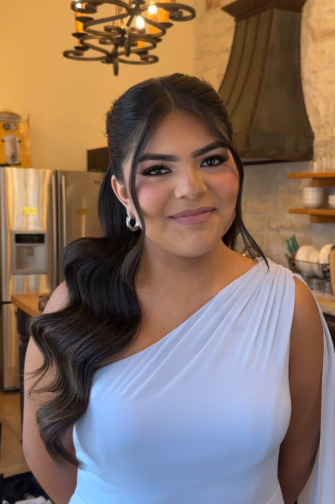 A woman with long, wavy dark hair, wearing a white dress, standing in a kitchen with a warm ambiance, kitchen appliances, and shelves in the background.