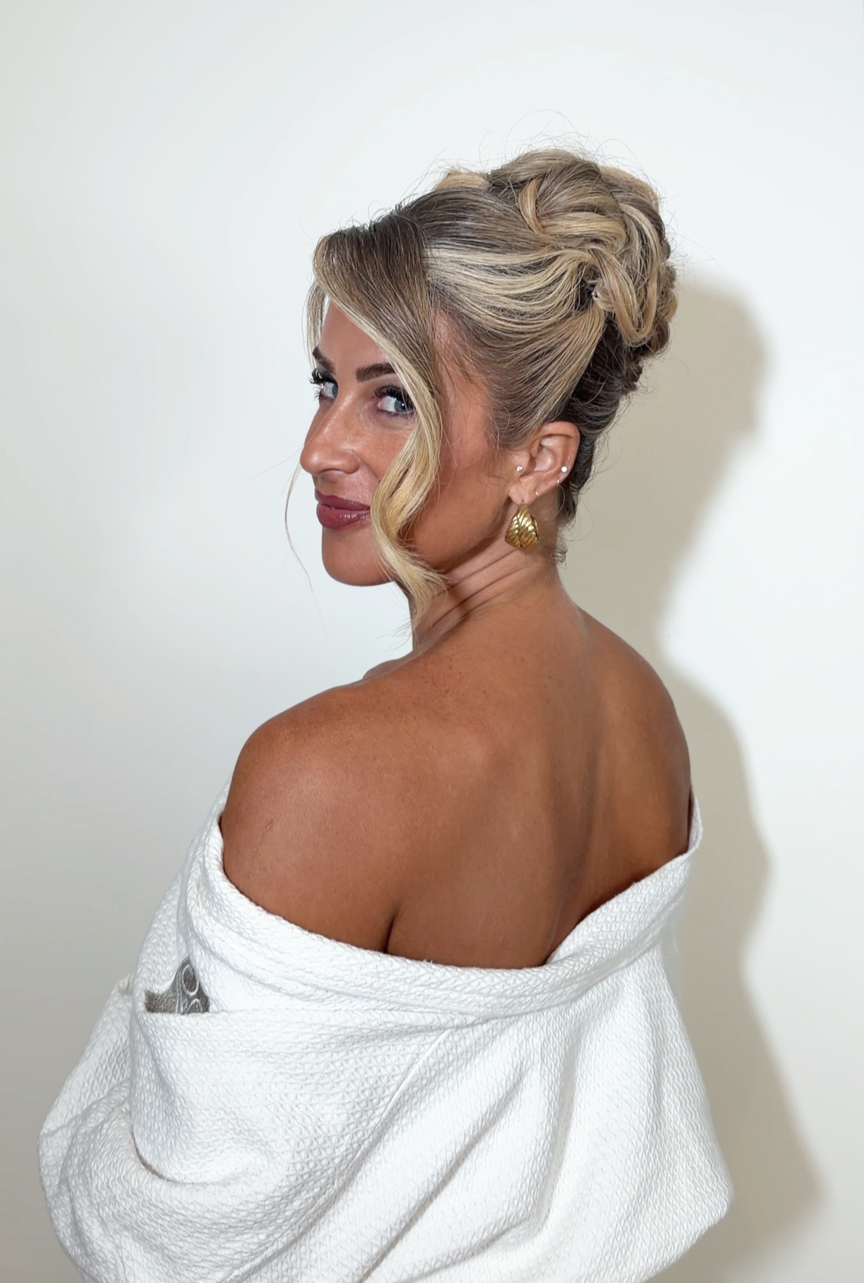 A woman with styled blonde hair in an elegant updo, wearing gold earrings and a white textured off-shoulder top, posed against a plain light-colored background.