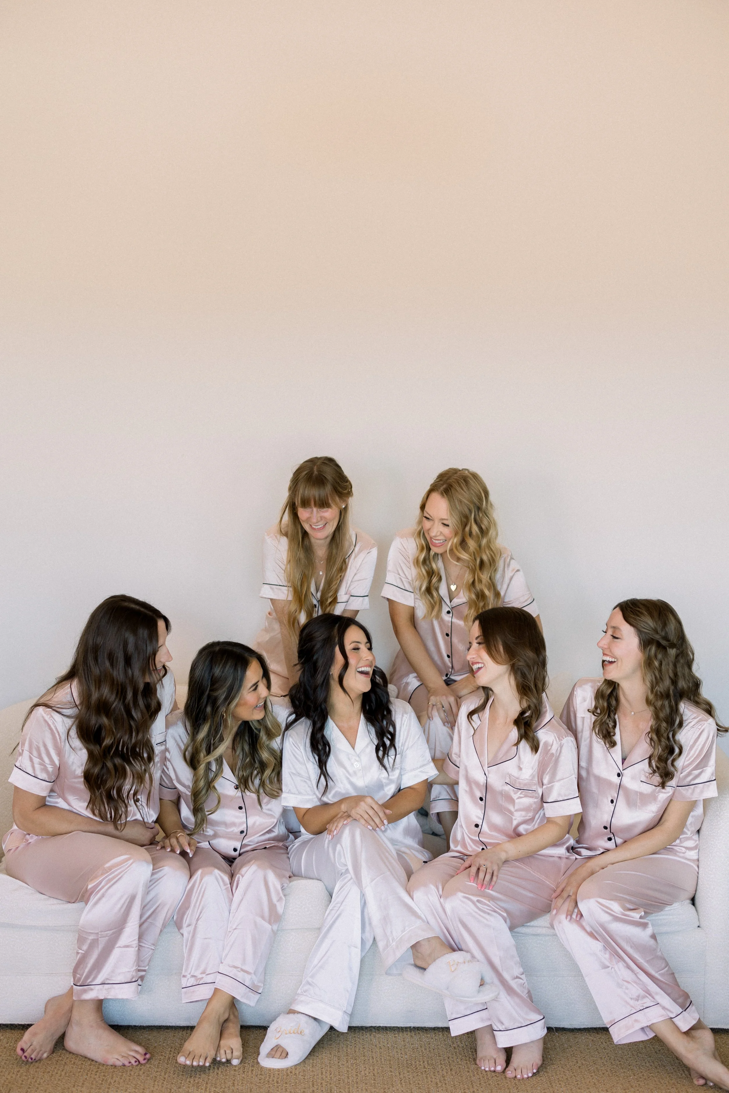 Group of women in matching pink pajamas sitting on a white sofa, smiling and talking, in a casual indoor setting.