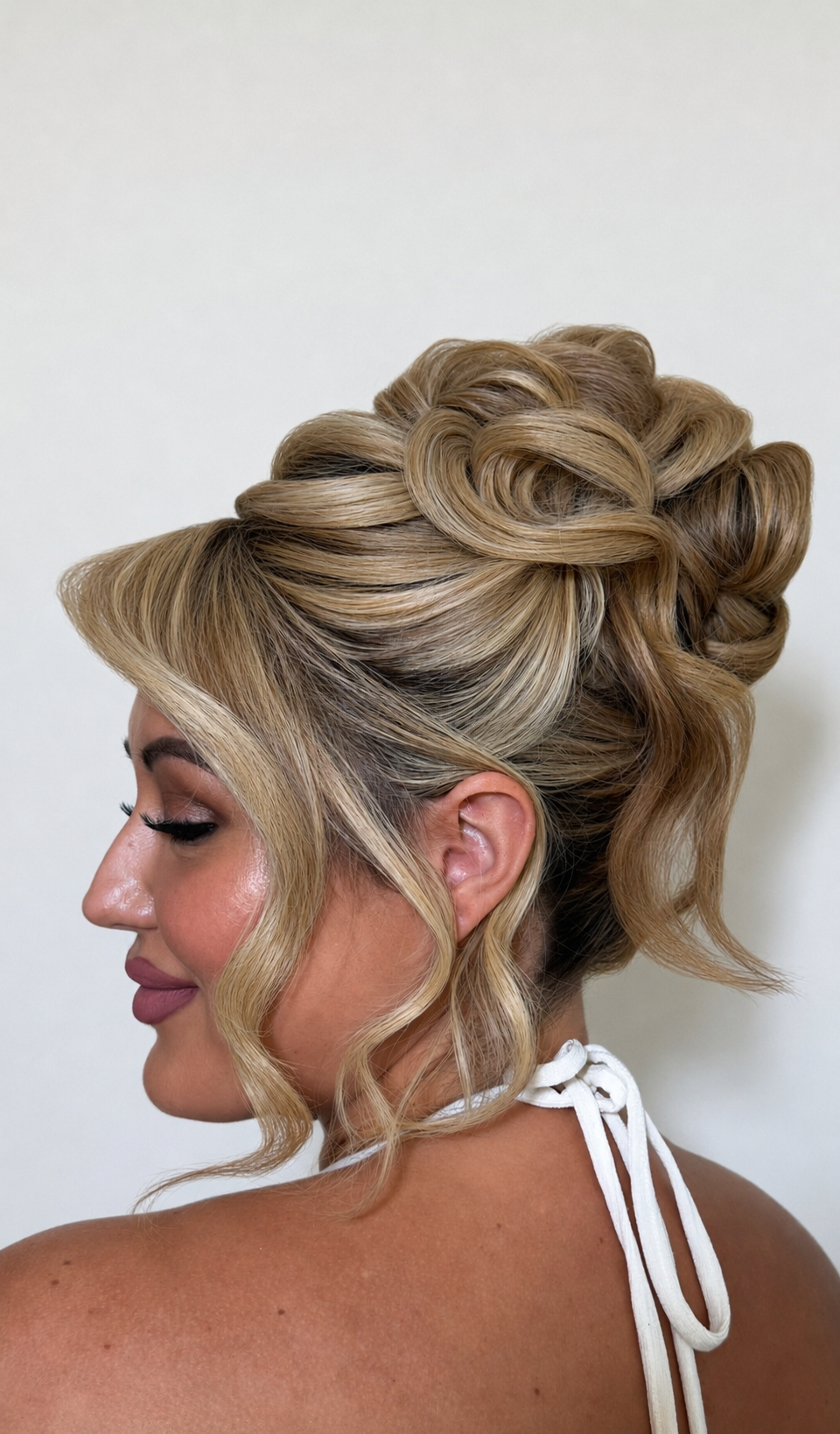 A woman with styled blonde hair in an elegant updo with soft curls and twists, wearing a white top with tie straps.