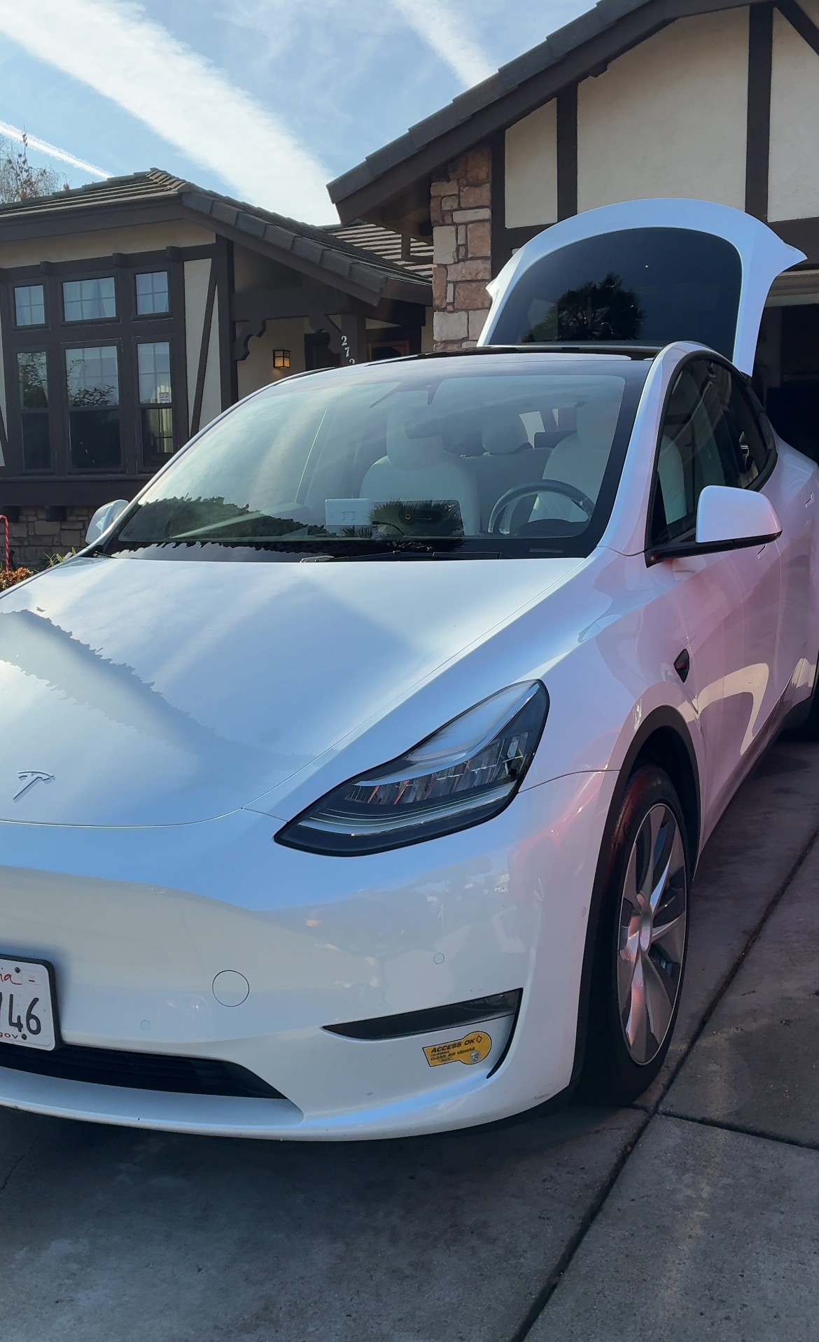 A white Tesla Model 3 with open front trunk parked in front of a house with a gray roof and brick and wood siding, under a blue sky.