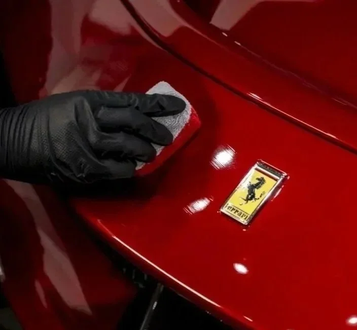 Person wearing black gloves polishing a red Ferrari logo on the car's surface with a cloth.