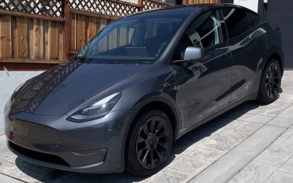 Gray Tesla Model 3 parked on a light-colored tiled driveway with a wooden fence in the background.
