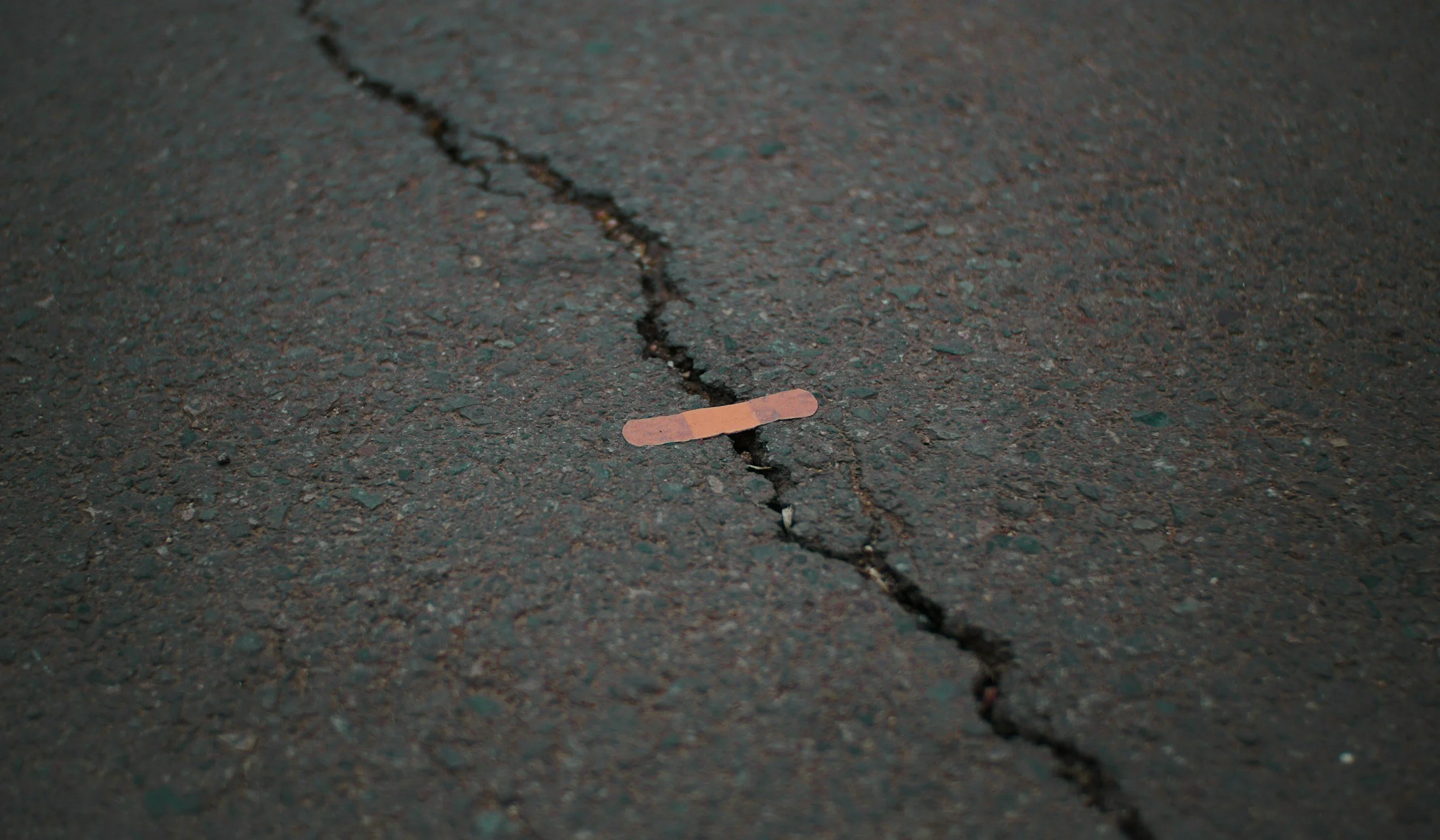 A cracked asphalt surface with a small Band-Aid placed over the crack.