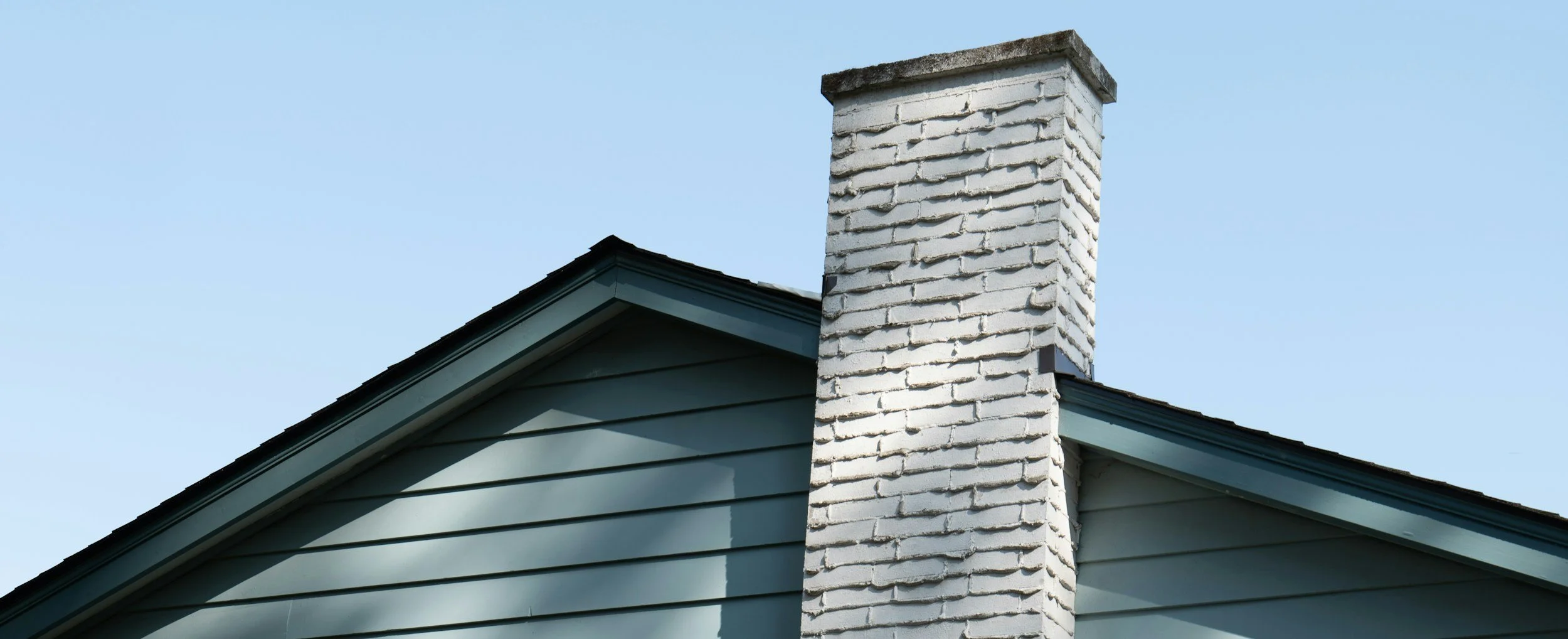 Close-up view of a house roof with a white brick chimney against a clear blue sky.