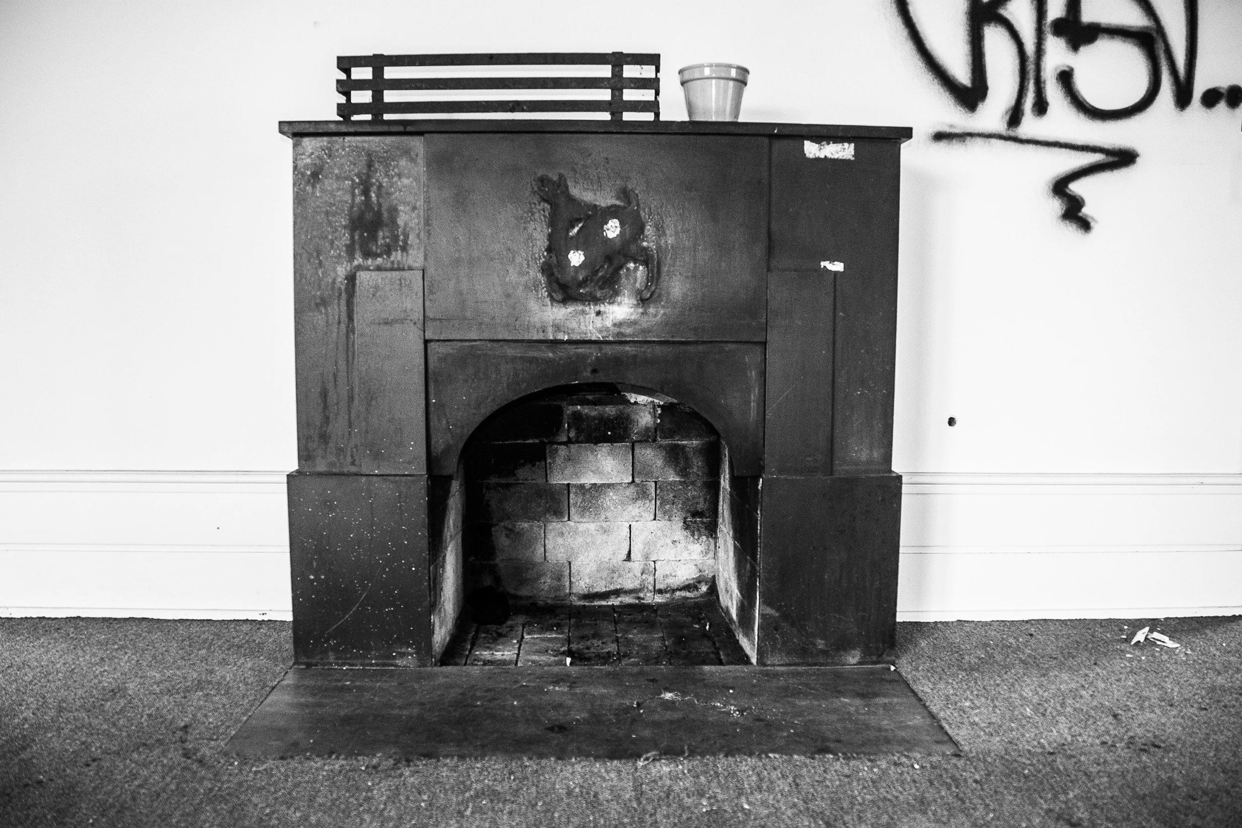 Black and white image of an old, rustic fireplace with a small wooden shelf on top holding a decorative pot. The wall next to the fireplace has graffiti. The floor is carpeted.
