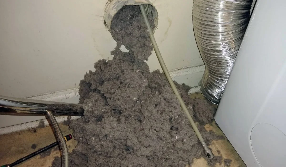A hole in the wall with insulation spilling out, next to a flexible metal duct, with a drain pipe and a washing machine nearby.