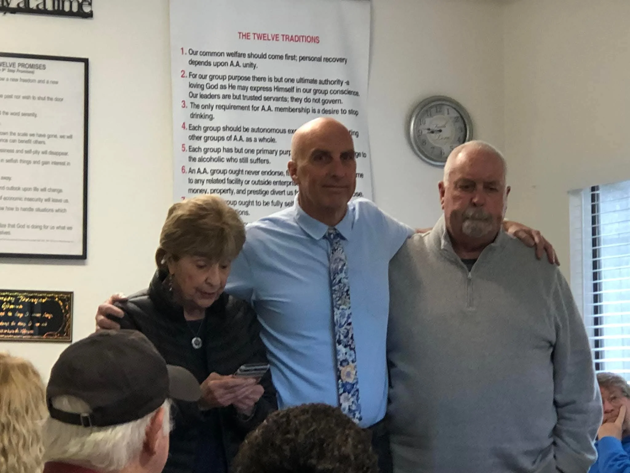 A group of people standing close together in a room with white walls, bulletin boards, and clock. Three individuals are in the foreground, with one woman holding a phone, and two men beside her. Others are partially visible in the background.