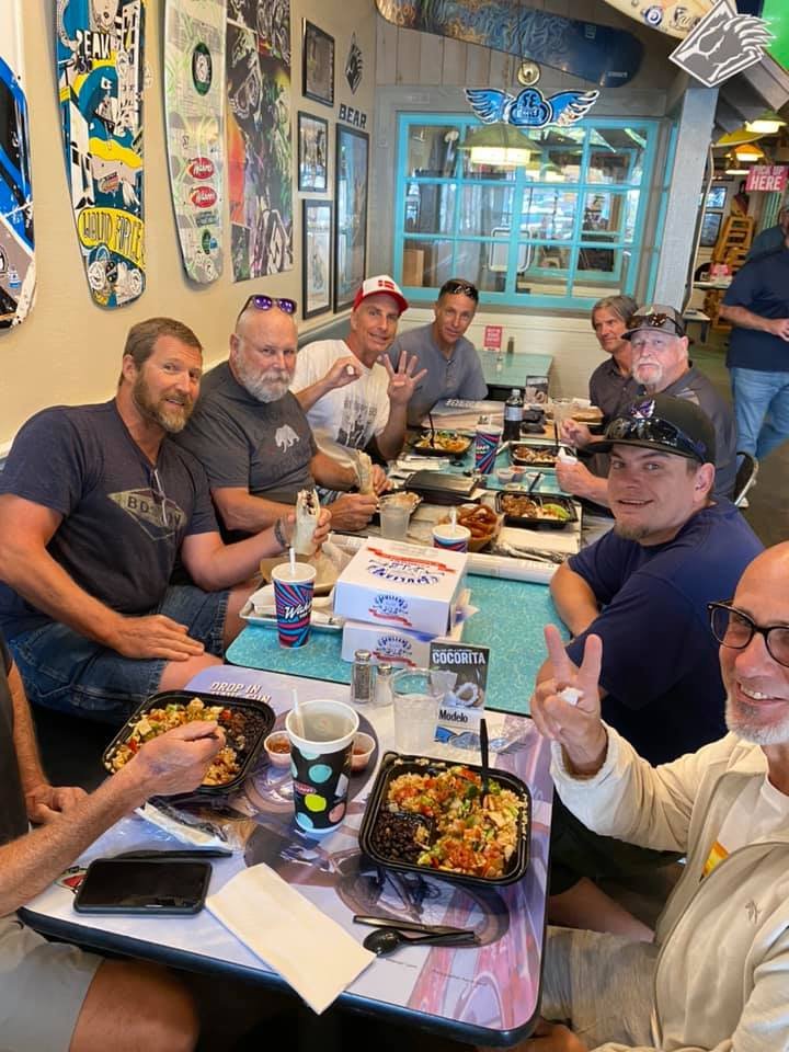 Group of men sitting at a restaurant table, eating and enjoying each other's company, with tacos and drinks on the table.