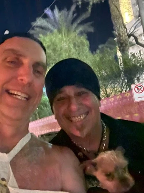 Two men smiling and posing for a selfie at night with a small dog. One man has short hair and the other is wearing a black beanie. There are trees and street signs in the background.