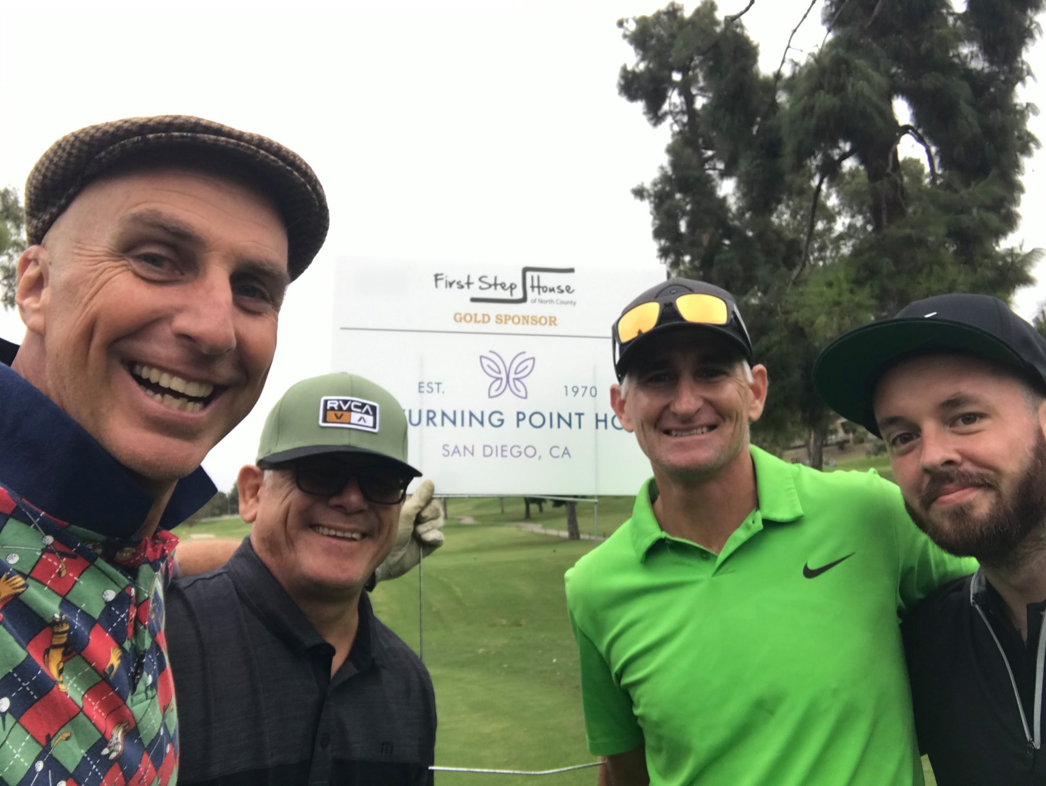 A group of five men taking a selfie outdoors at a golf course with trees in the background, standing in front of a sign that indicates a location and sponsorship details for an event in San Diego, California.