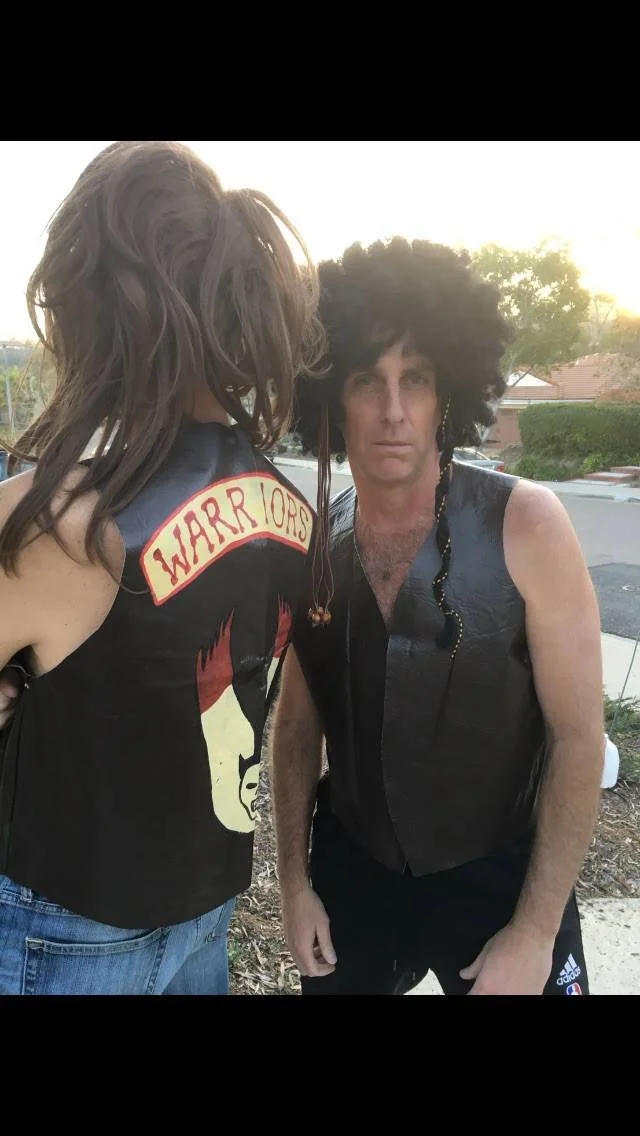 Two men dressed in rock-inspired costumes posing outdoors during daytime. The man on the right has a large curly wig, sleeveless leather vest, and is making a serious face. The woman on the left, whose face is not visible, has long brown hair, a vest
