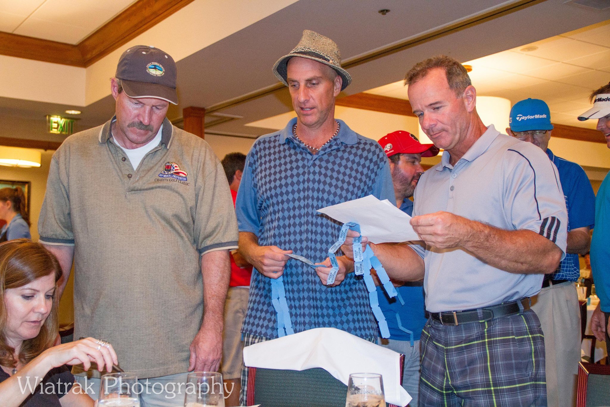 Four men in casual clothing, two wearing golf caps, are examining documents and raffle tickets at a social event in a hotel banquet room. One man is holding raffle tickets, another seems to be reading a paper, and the others look on.