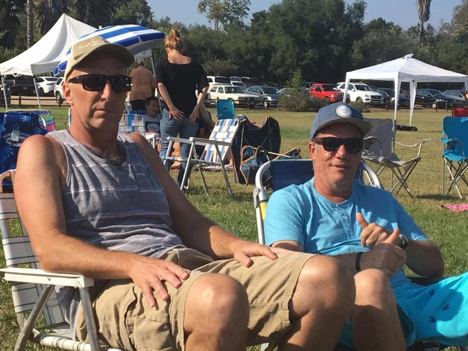 Two men sitting outdoors in lawn chairs at a gathering, with tents and other people in the background, on a sunny day.