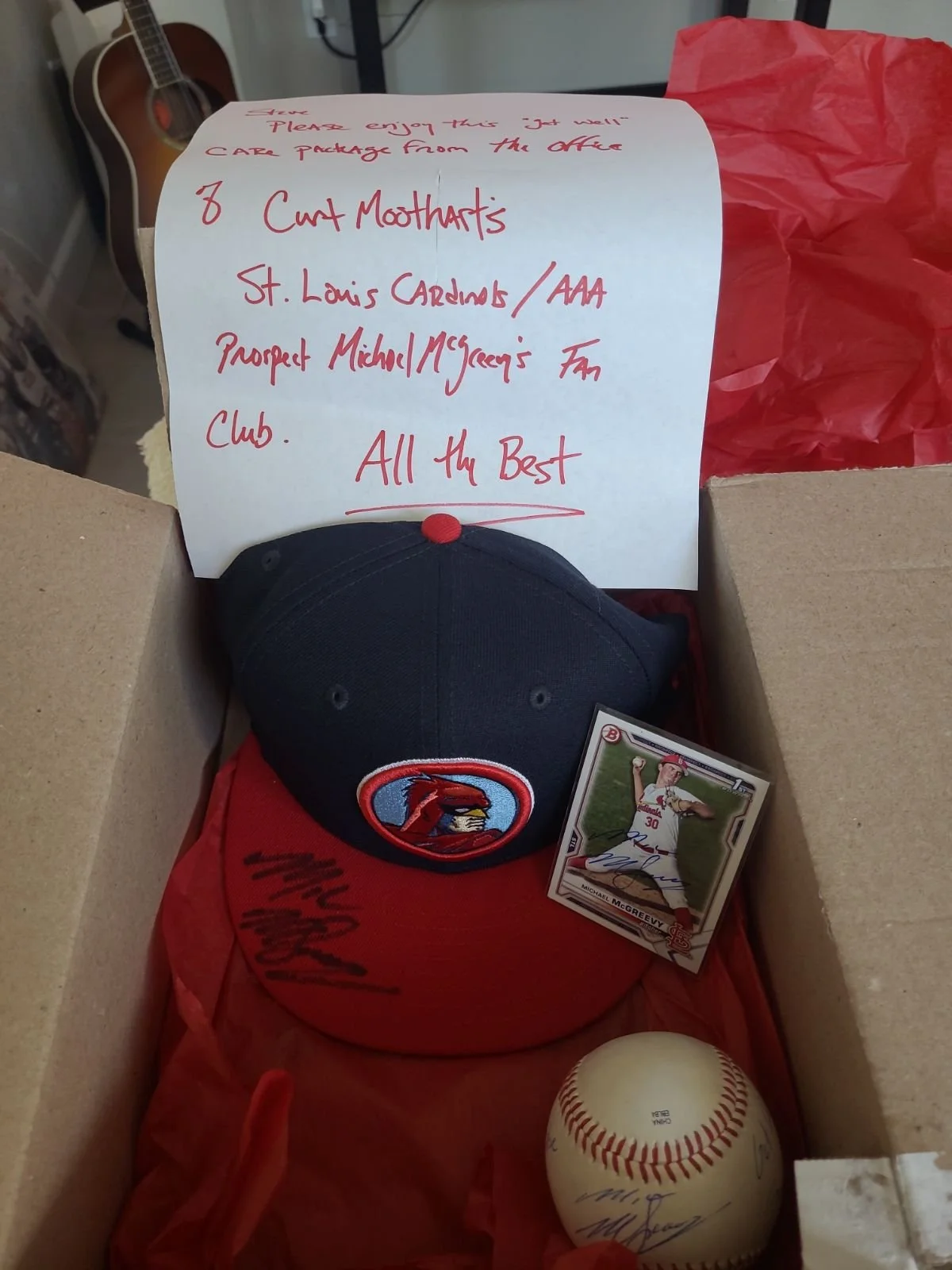 A cardboard box containing a signed baseball, a black and red baseball cap with an Arizona Diamondbacks logo, a photograph of a baseball player (Michael McGreevy) with his autograph, and a handwritten note on white paper mentioning donations to a cha