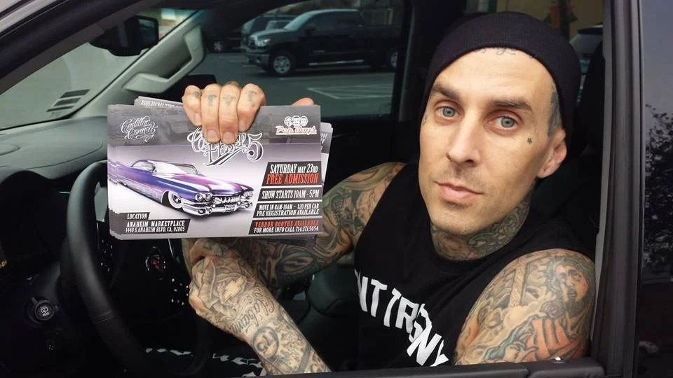 Tattooed man sitting in the driver's seat of a vehicle, holding a flyer for a car event, with a piercing in his right eyebrow, wearing a black head covering and a black shirt.