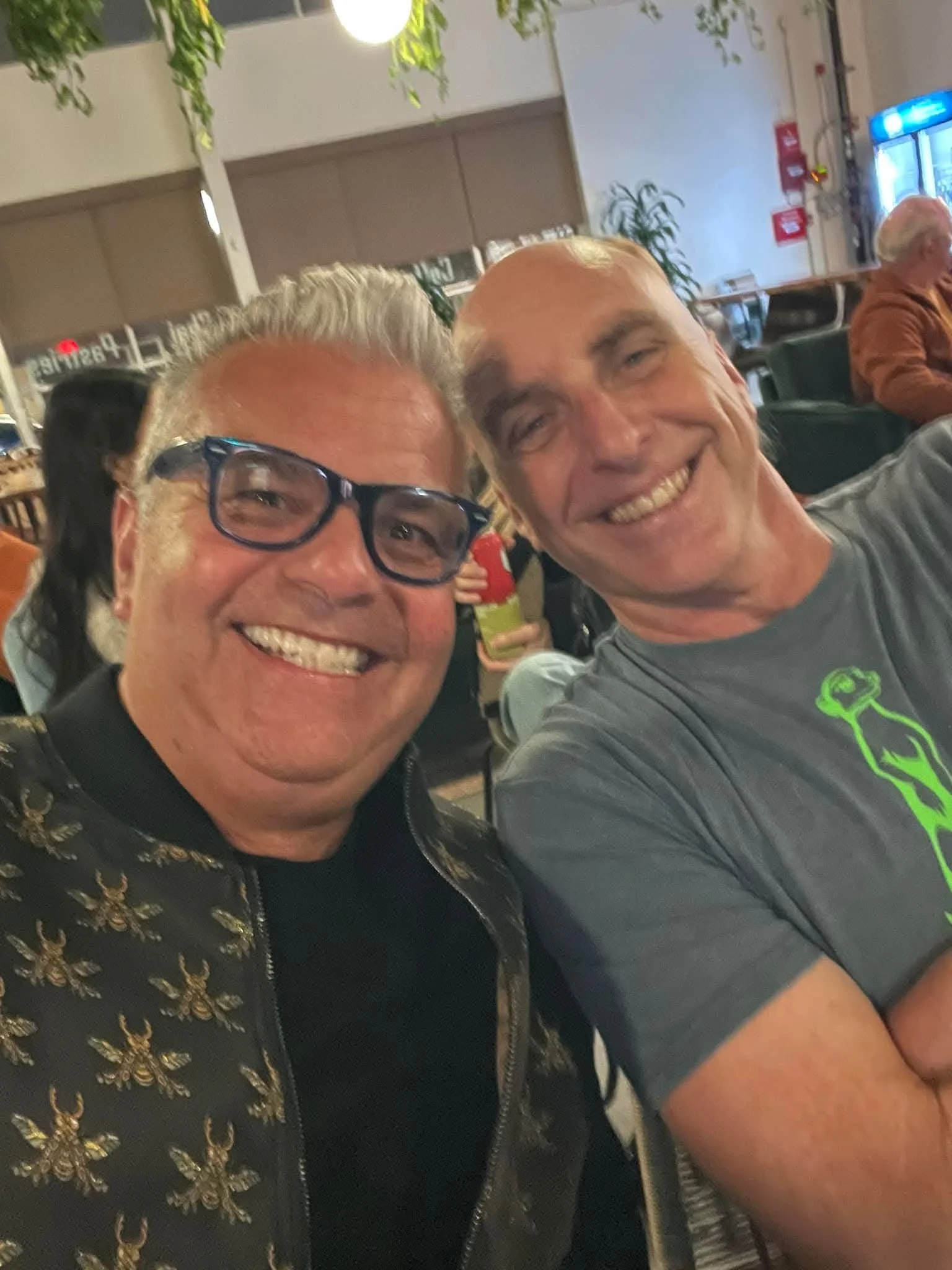 Two older men smiling for a photo in a social setting, with other people and hotel or restaurant decor in the background.