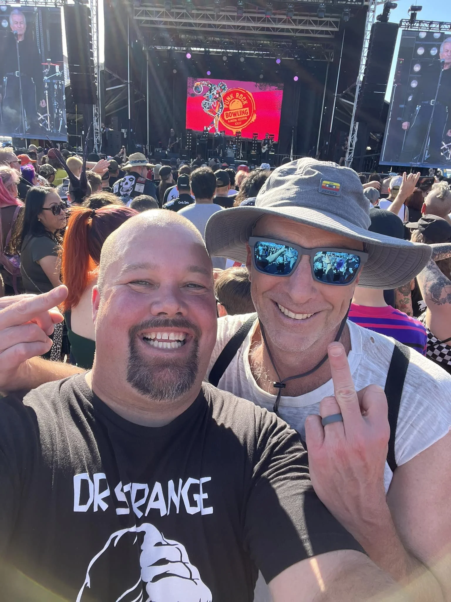 Two smiling men taking a selfie at a crowded outdoor music festival. The background features a large stage with a screen displaying a pink and red logo for the Punk Rock Bowling festival. The crowd includes people of various ages, some wearing hats a