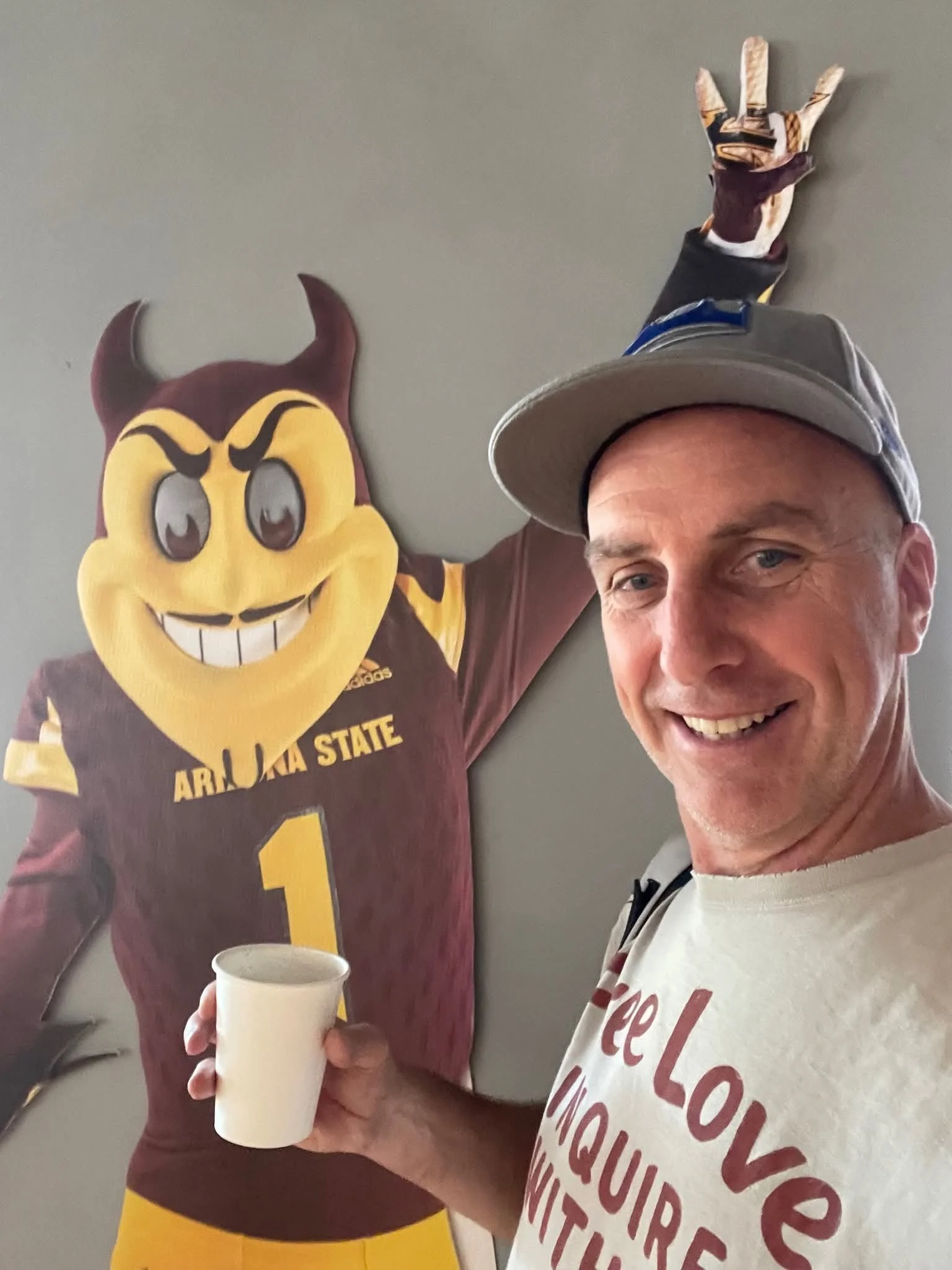 A man smiling, wearing a gray cap and a white T-shirt with red text, holding a white cup, posing next to a person dressed in a mascot costume resembling a yellow bird with a brown body and a big head with angry eyebrows, large eyes, and a wide grin w
