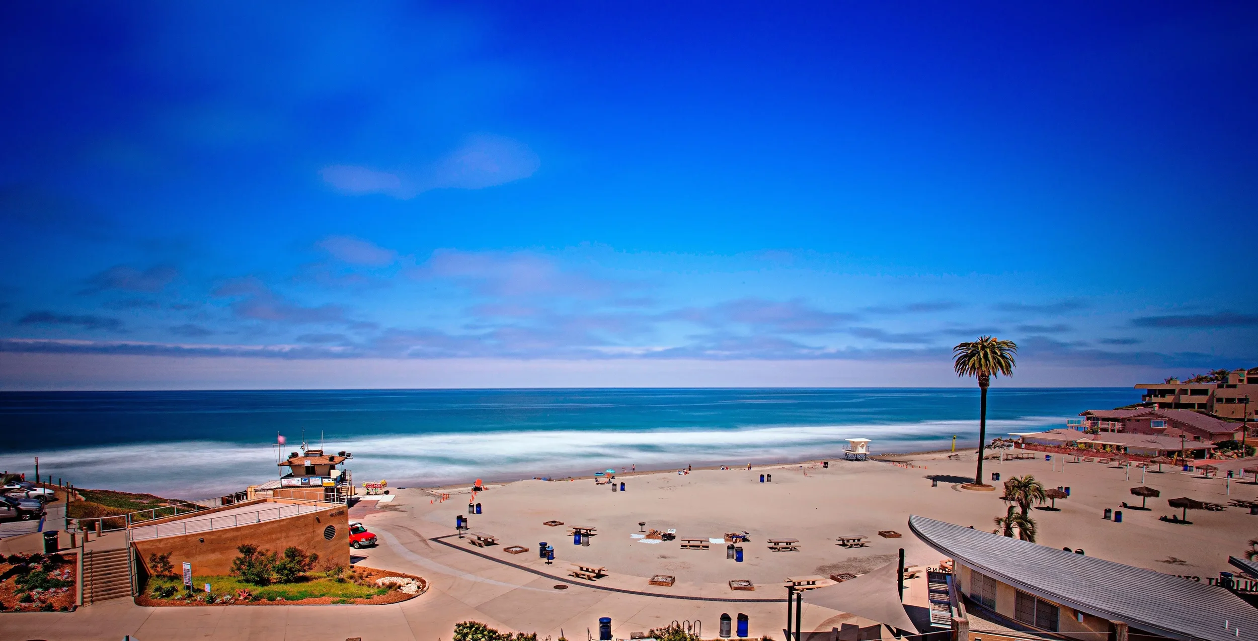 A sunny beach scene with a palm tree, lifeguard tower, benches, umbrellas, and a few buildings along the shoreline, with a vast blue sky and calm ocean waves.