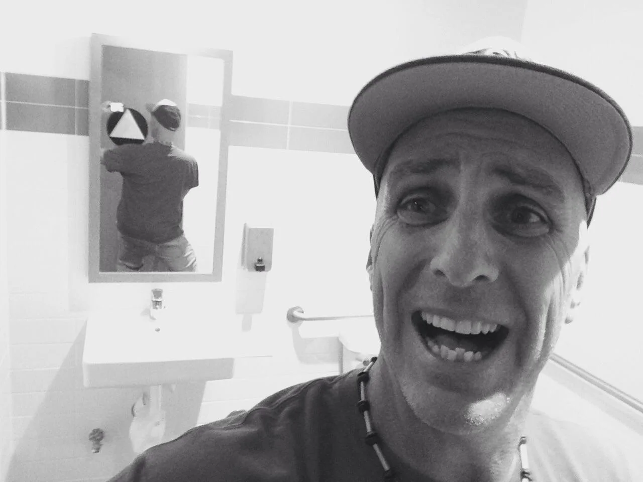 A man smiling and taking a selfie in a bathroom with a mirror behind him showing his back, wearing a baseball cap and necklace.