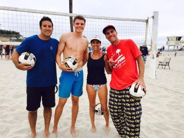 Four people standing on a sandy beach volleyball court, holding volleyballs, with a net behind them and beachgoers in the background.