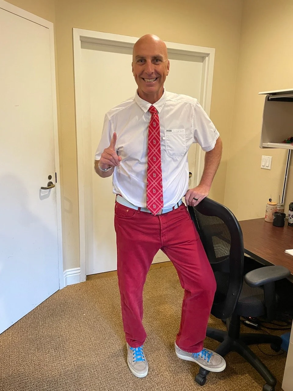 A bald man wearing a white dress shirt with rolled-up sleeves, a red patterned tie, bright red pants, and beige sneakers with blue laces standing in an office, smiling, and holding up one finger.
