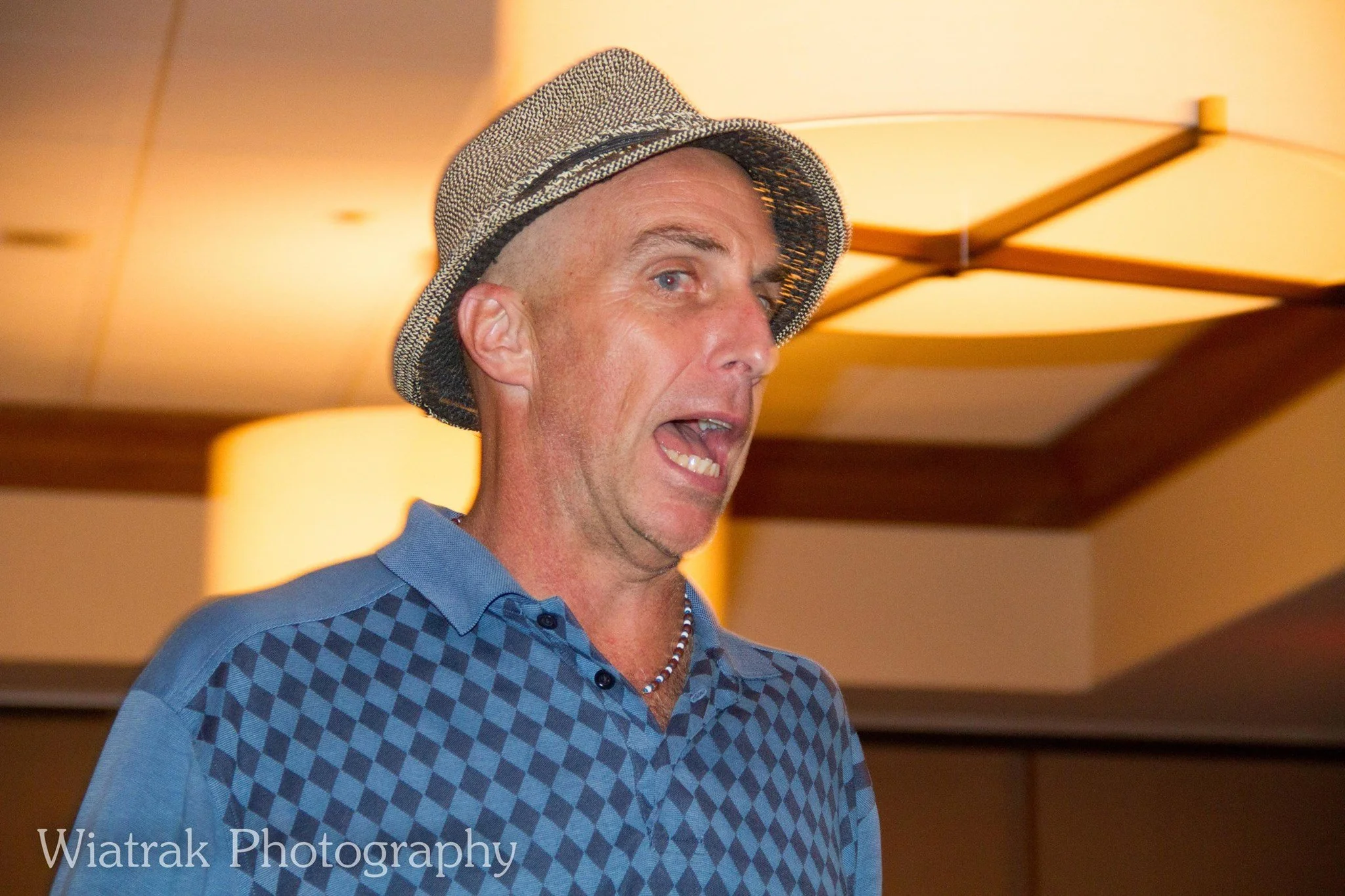 A man wearing a gray hat and a blue shirt with a diamond pattern, speaking or singing with his mouth open.