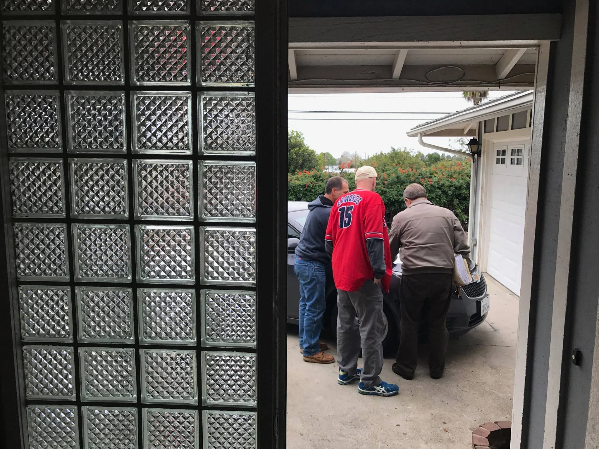 Four people gathered outside a garage, seen through a doorway with glass blocks on the left and a white garage door with windows on the right. The scene shows a suburban neighborhood with trees and power lines in the background.