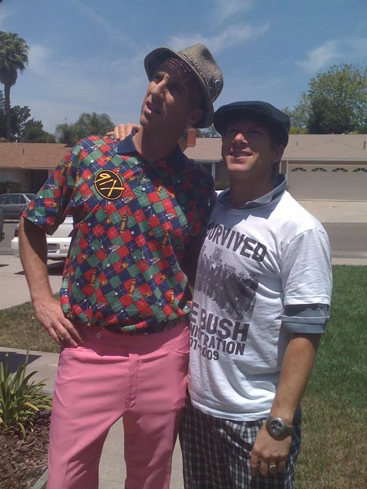Two men standing outside on a sunny day, one wearing a colorful plaid shirt, pink pants, and a hat, and the other wearing a white political t-shirt and plaid shorts, smiling and posing for a photo.