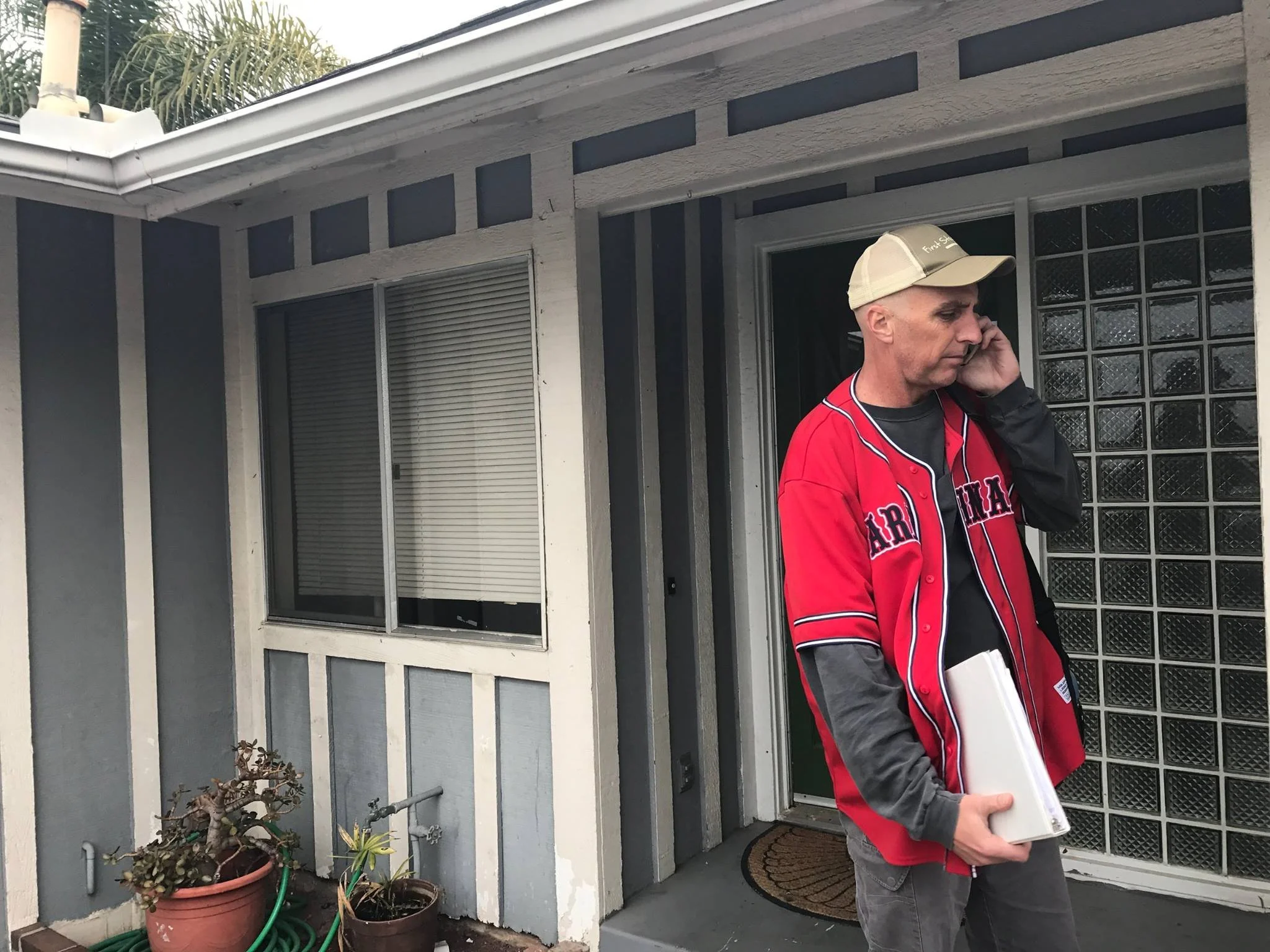 A man wearing a beige cap and a red and black sports jacket is talking on a cellphone while holding a book or folder outside a house with gray siding and glass block windows. There are potted plants and a garden hose near the house.