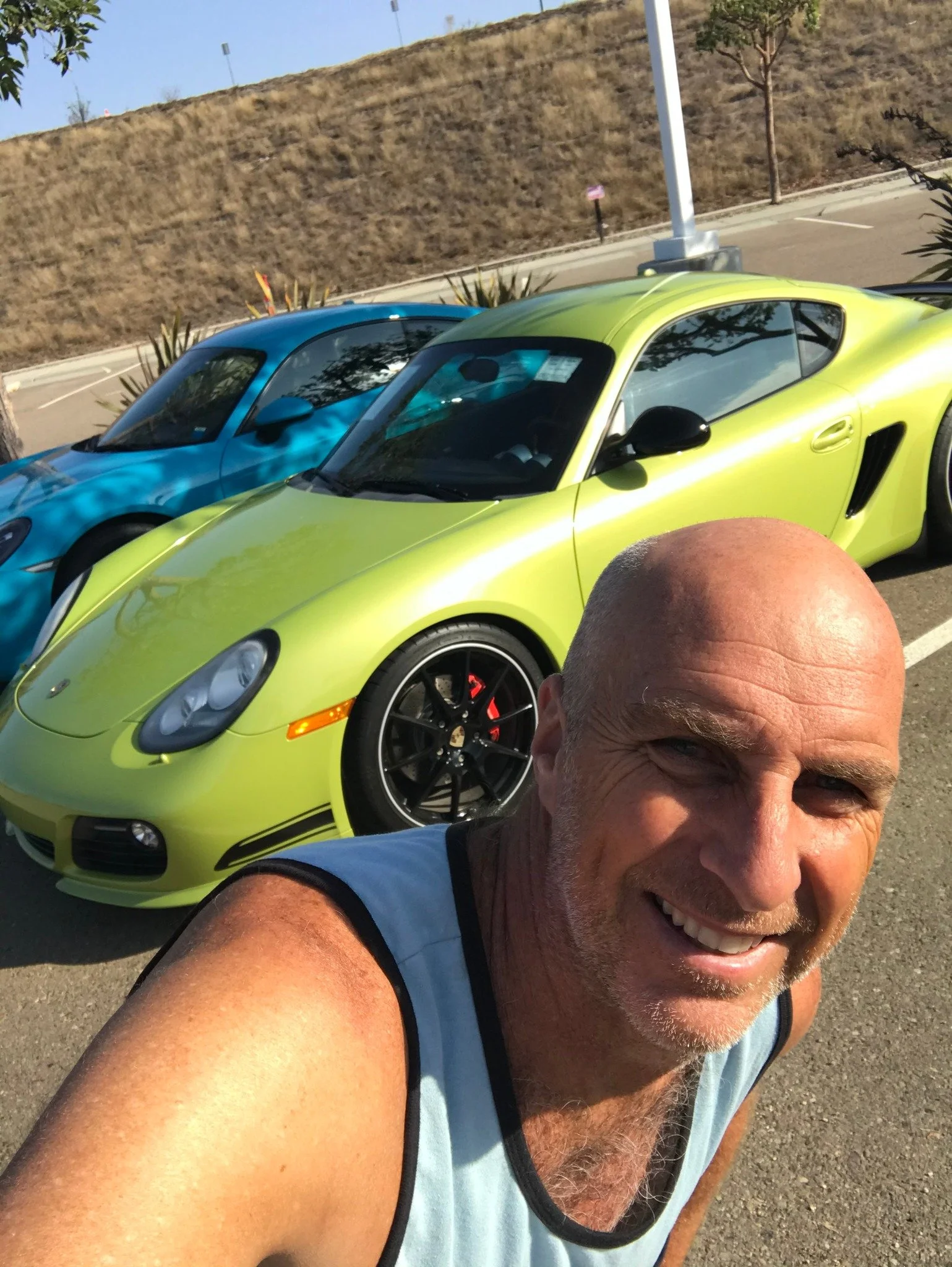 A smiling man with a shaved head taking a selfie in front of two sports cars, a green one and a blue one, parked outdoors. The background includes a dry hillside, parking lot, and some trees.