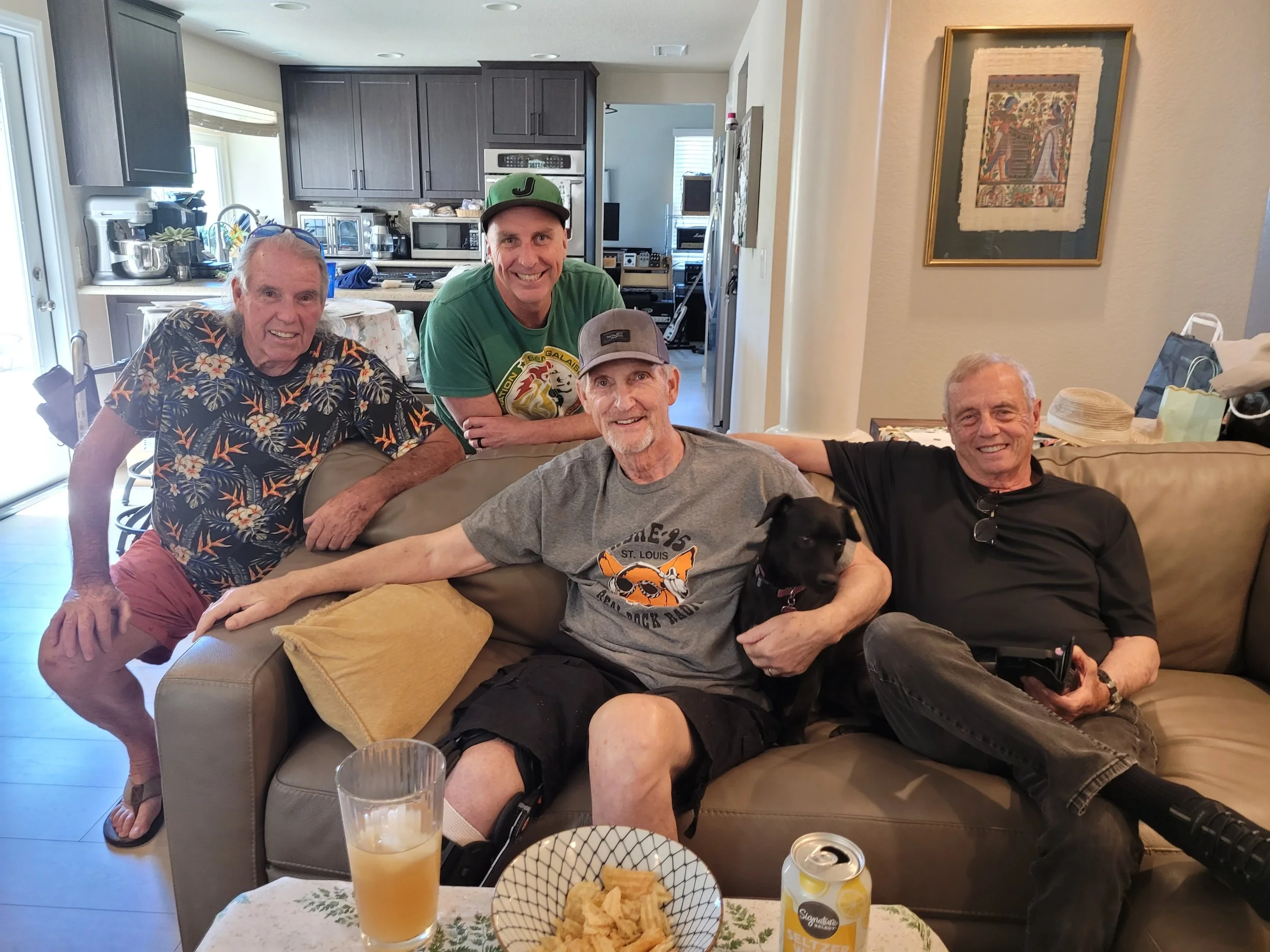 Group of five men sitting and standing in a living room, smiling. There is a framed picture on the wall, a table with snacks and drinks, and kitchen cabinets in the background.