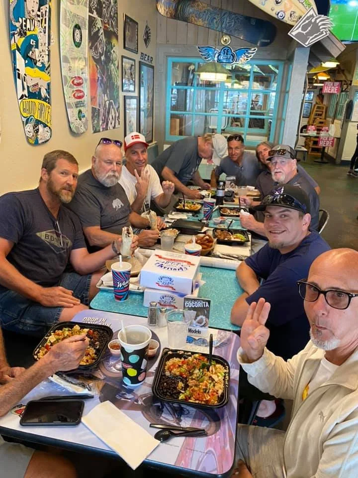 A group of nine men sitting around a table at a restaurant, enjoying a meal together. The table has food containers, drinks, and utensils. The restaurant has decorated walls with skateboard art and posters, and surfing flags are visible hanging from 