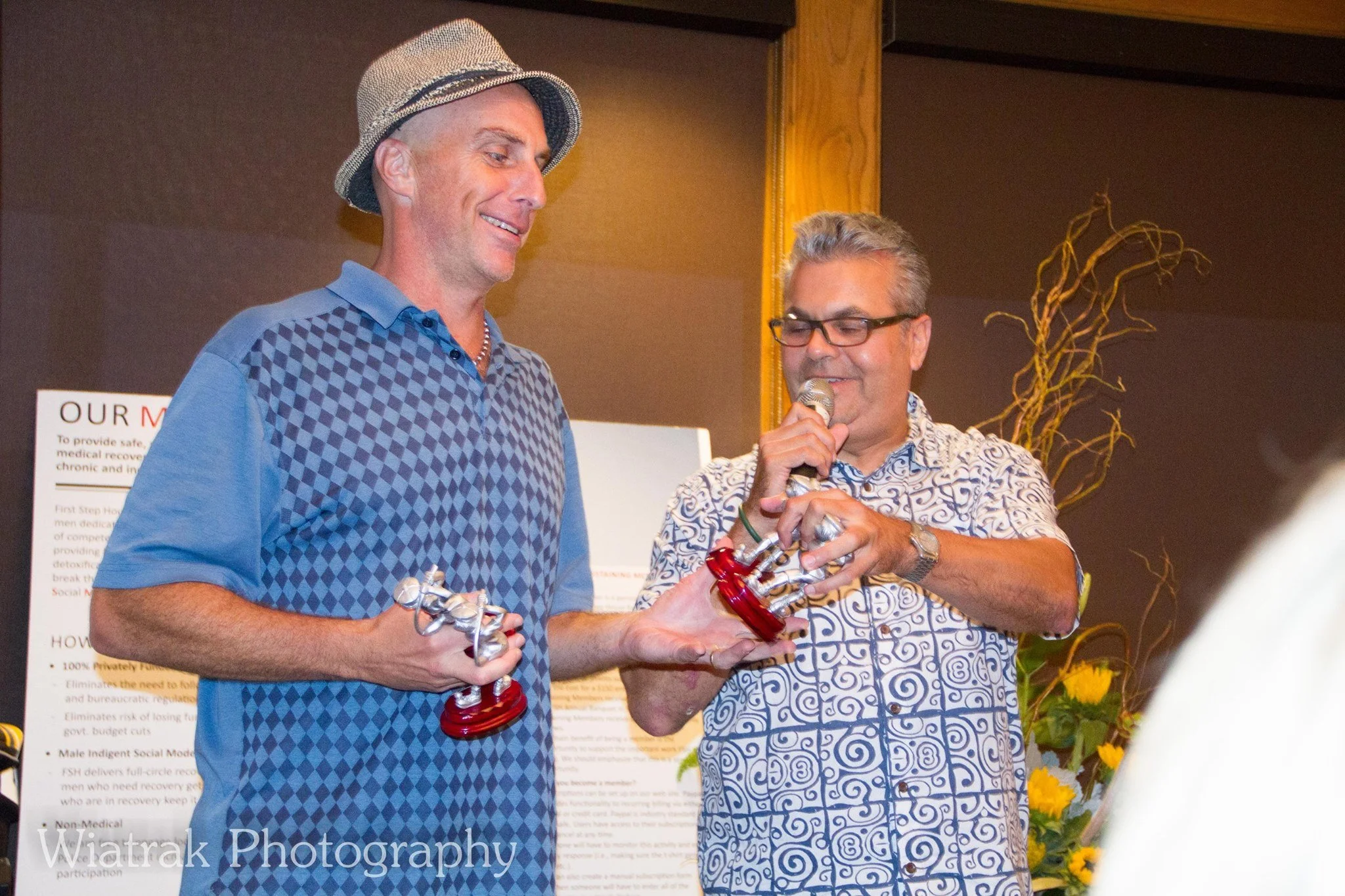 Two men are standing together indoors, with one man seemingly presenting an award to the other. The man on the left is wearing a checkered blue shirt and a hat, while the man on the right is wearing glasses and a patterned shirt, holding a microphone