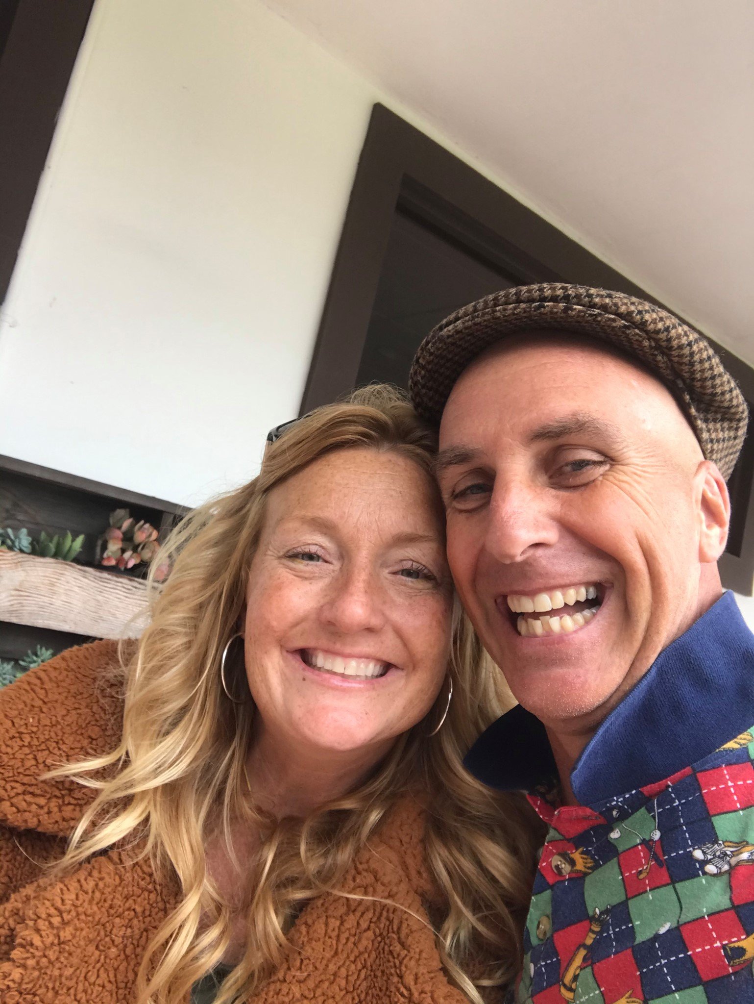 Close-up of two people smiling, a woman with long blonde hair and a man wearing a patterned hat and a colorful shirt, indoors in front of a white wall and a dark door frame.
