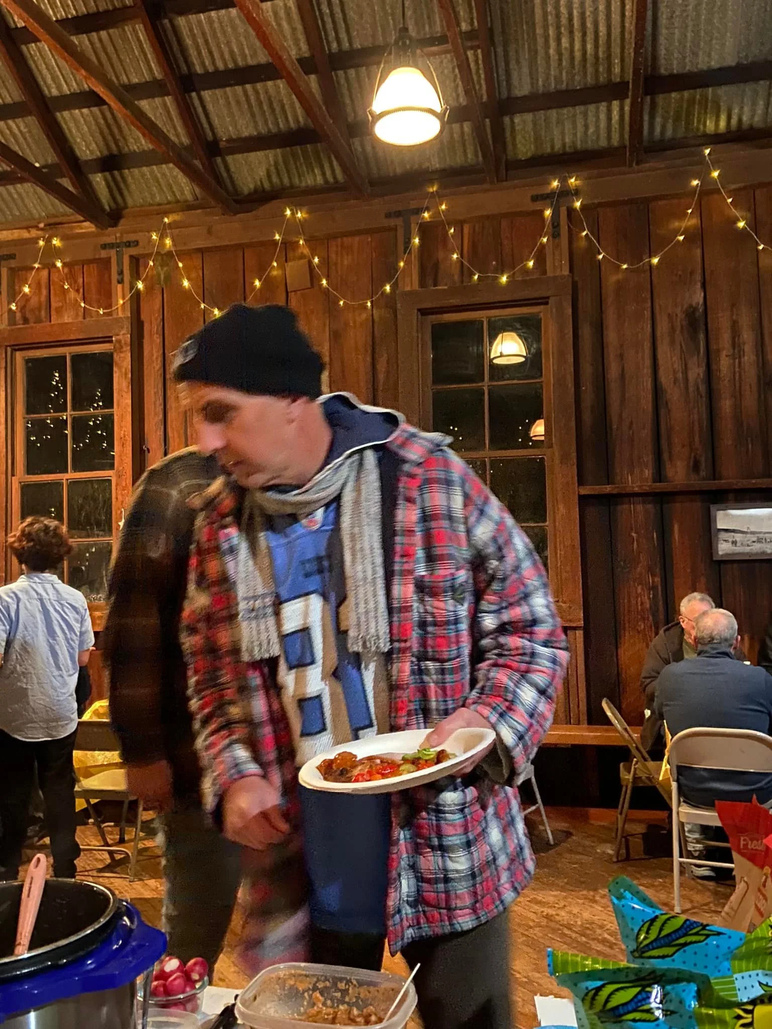 A man in a plaid shirt and scarf holds a plate of food in a rustic indoor setting, with other people and warm lighting in the background.