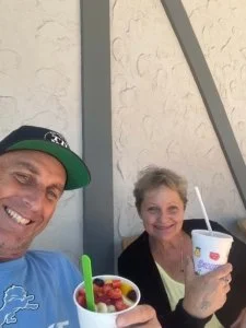 A man and an elderly woman smiling and holding cups of frozen yogurt, outdoors against a textured wall with vertical gray beams.