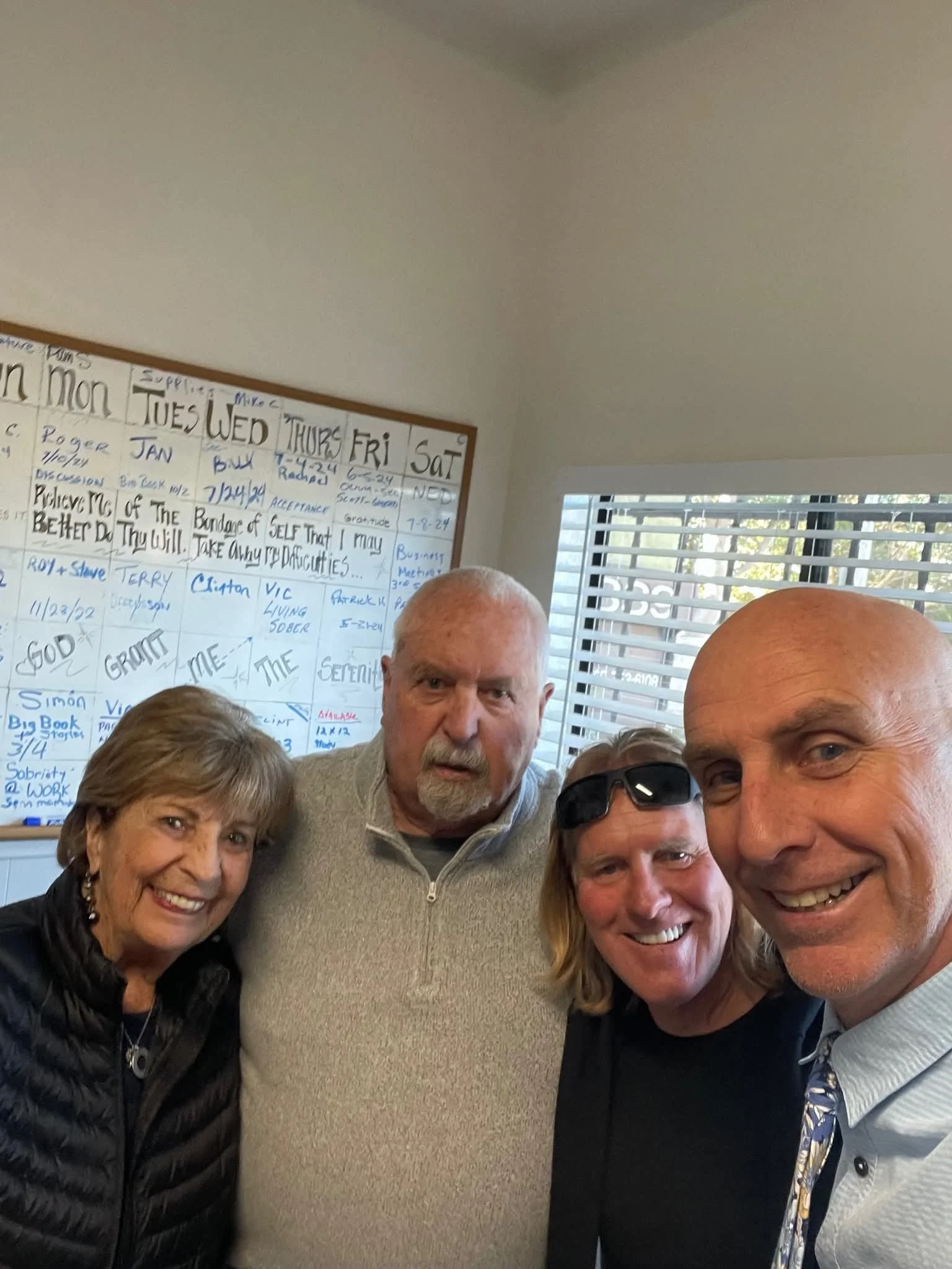 A group of five people, four men and one woman, smiling and taking a selfie indoors with a whiteboard and window in the background.