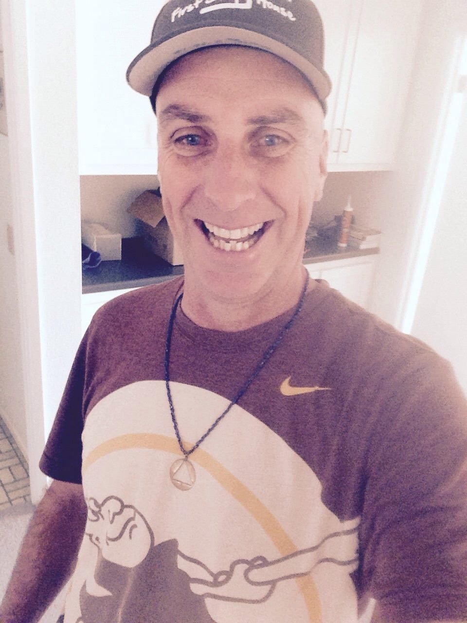 A man smiling and wearing a baseball cap, a graphic t-shirt, and a necklace, taking a selfie in a kitchen.