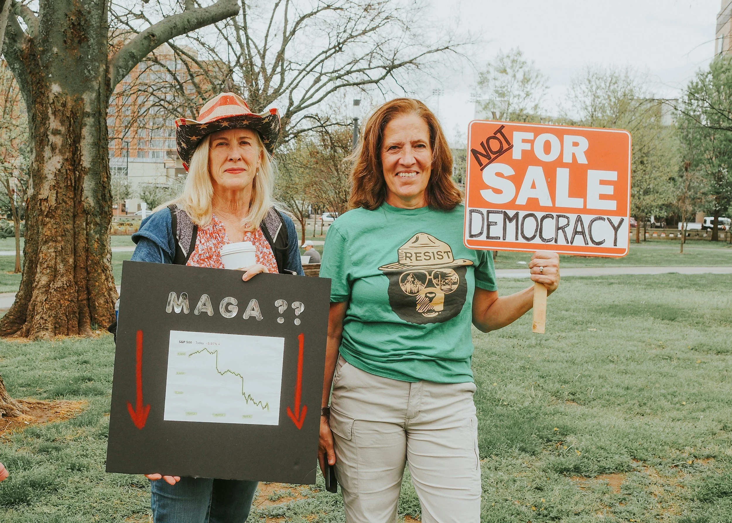 Two women standing outdoors in a park holding protest signs. The woman on the left holds a black sign with a declining stock chart and the text 'MAGA??'. The woman on the right holds an orange and white sign that reads 'NOT FOR SALE DEMOCRACY'. Both women appear serious, and there are trees and a cityscape in the background.