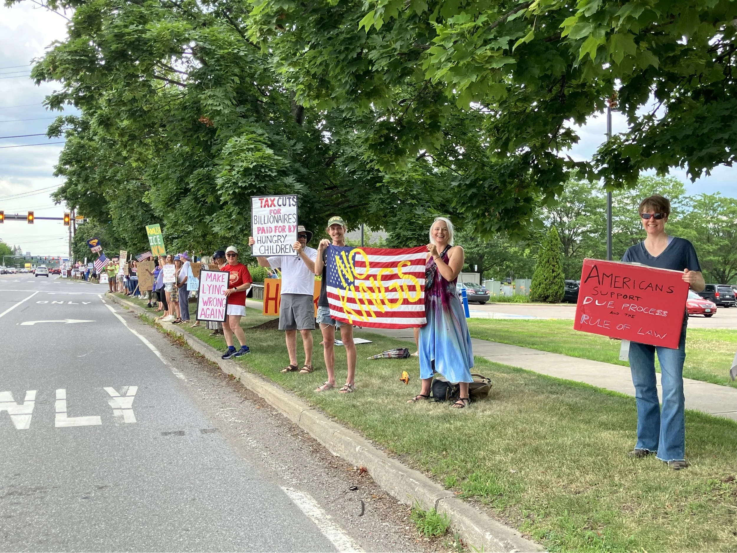 A group of protesters standing on the grass beside a street during a protest, holding various signs that express political opinions about tax cuts, government support, and legal processes.