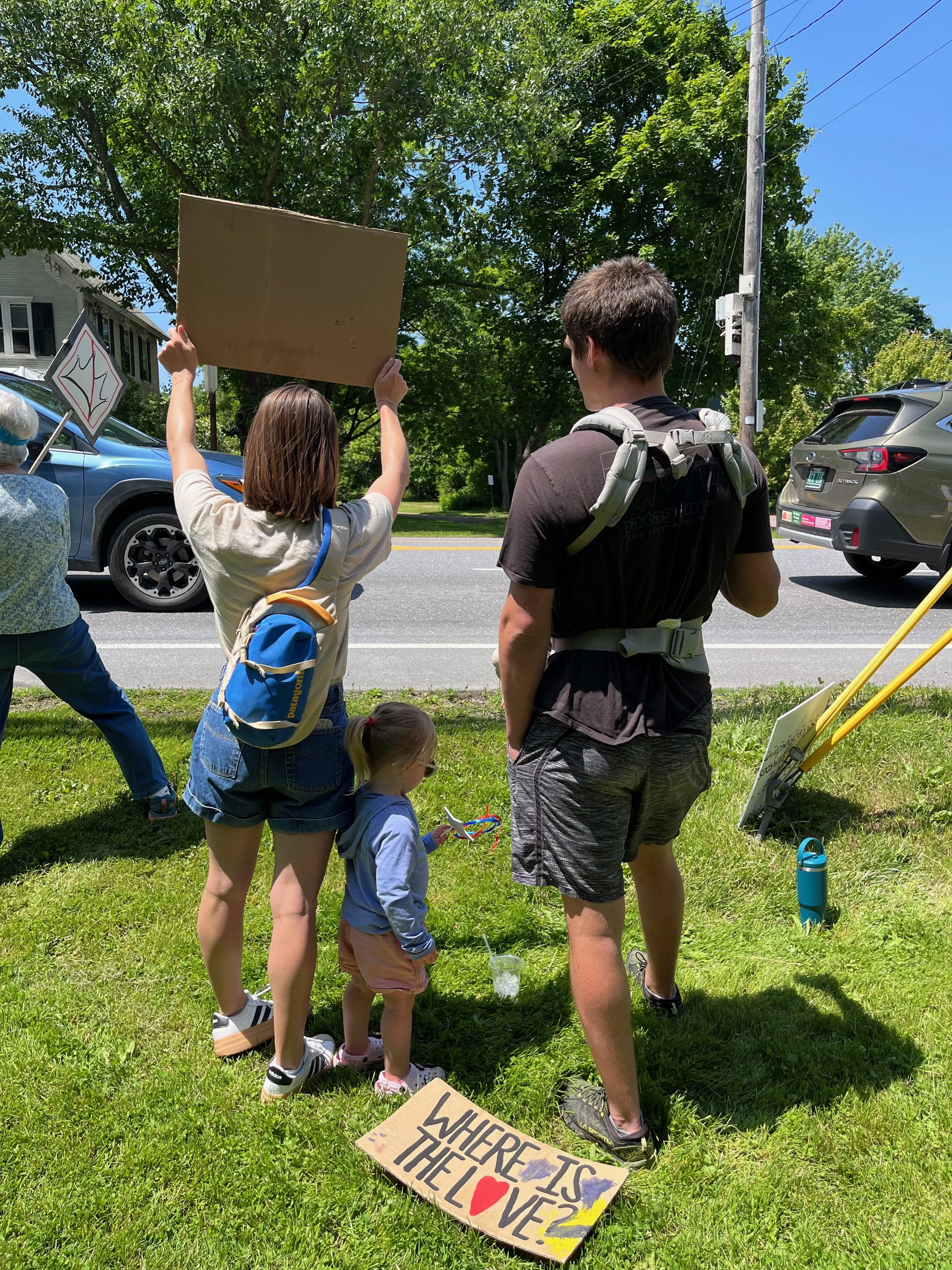 A group of people, including children, participating in a protest or demonstration on a grassy area next to a street. Some hold signs, with one visible sign reading "WHERE IS THE LOVE?" The scene is sunny with green trees, parked cars, and utility po