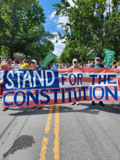 People participating in a parade or protest march holding a large banner that reads 'STAND FOR THE CONSTITUTION' with green signs in the background and trees lining the street.