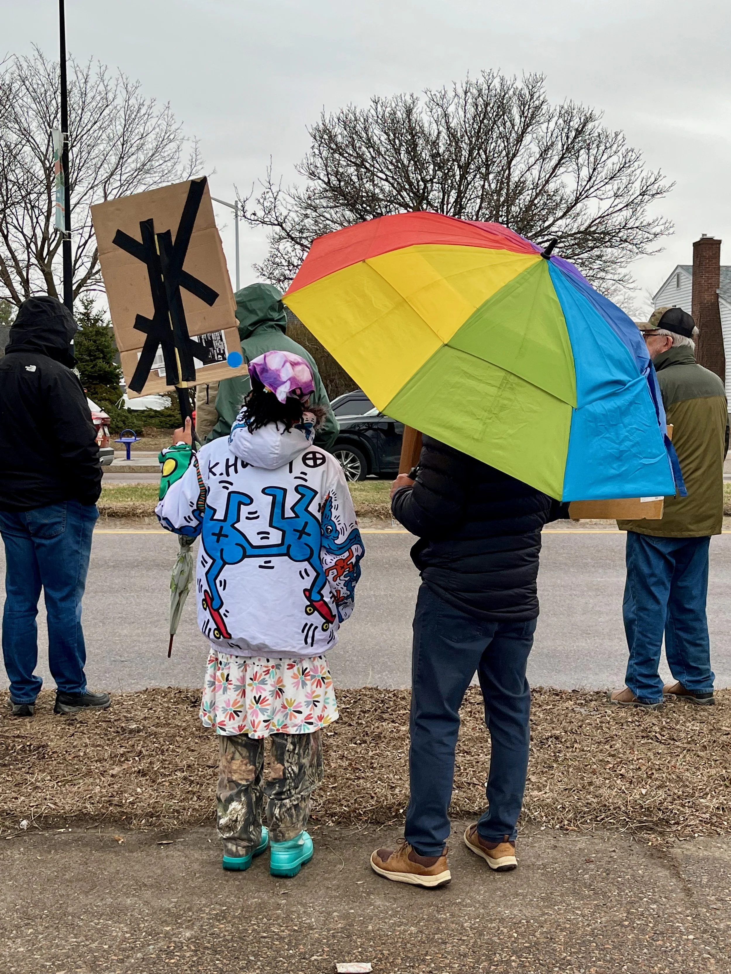 People protesting outdoors on a cloudy day, holding signs and umbrellas, with a woman in a colorful jacket and camouflage pants, and others in jackets and hats.